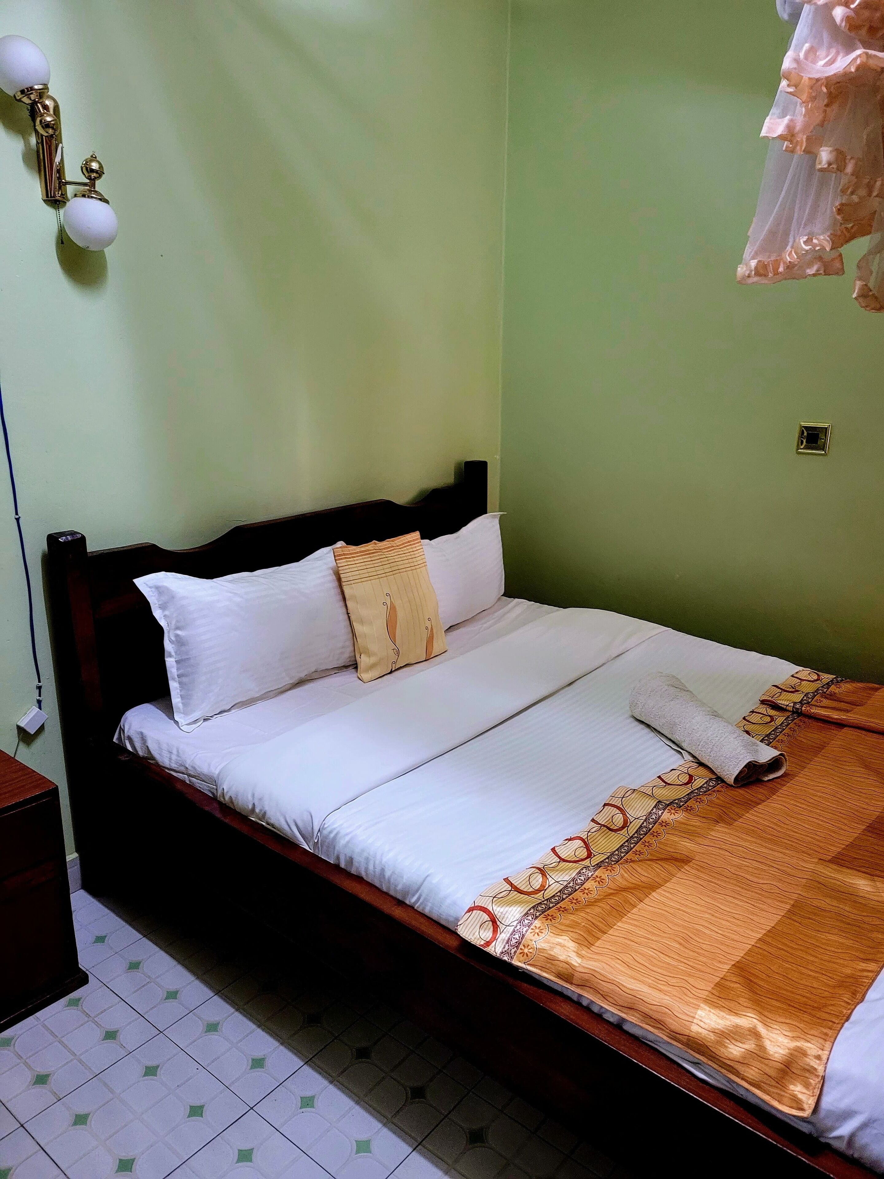A neatly made bed with white linens and a patterned blanket, a bedside table with a lamp, and a wall-mounted light fixture.