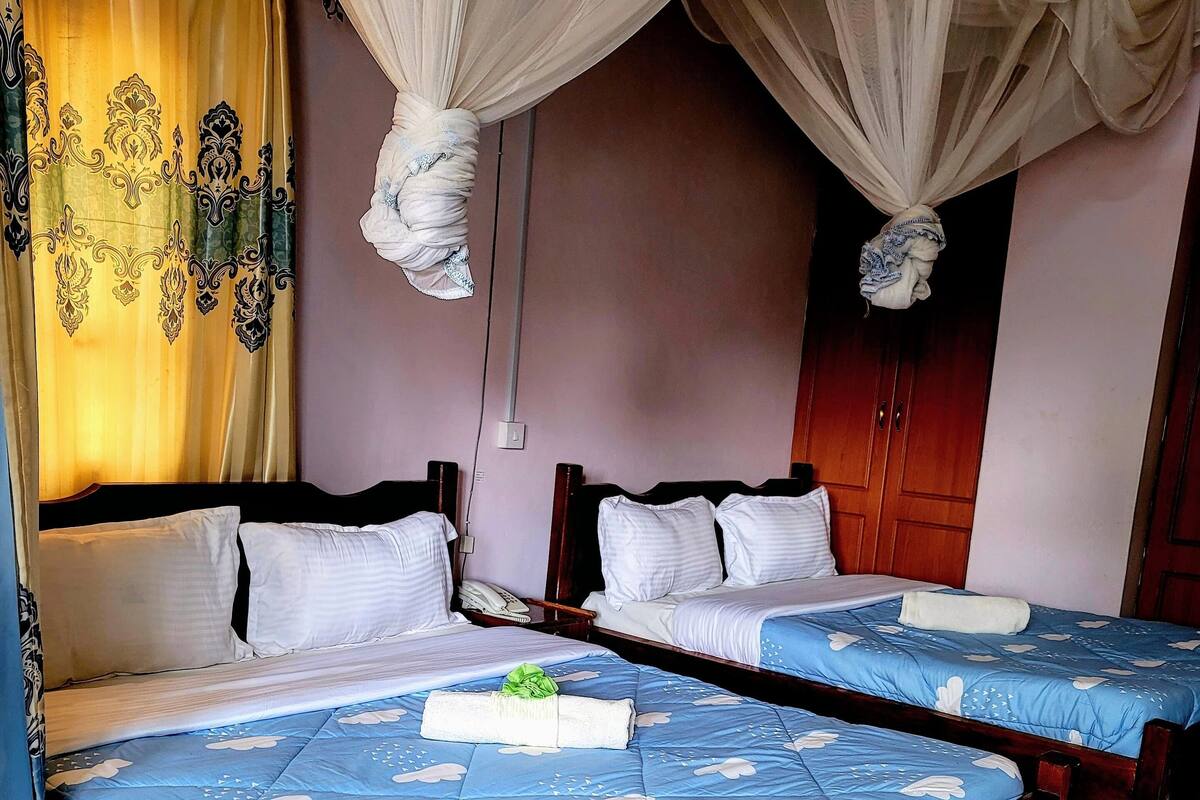 A room with two beds, each with a blue bedspread and white pillows. There is a curtain with a yellow and black pattern on one wall, and a wooden door in the background.