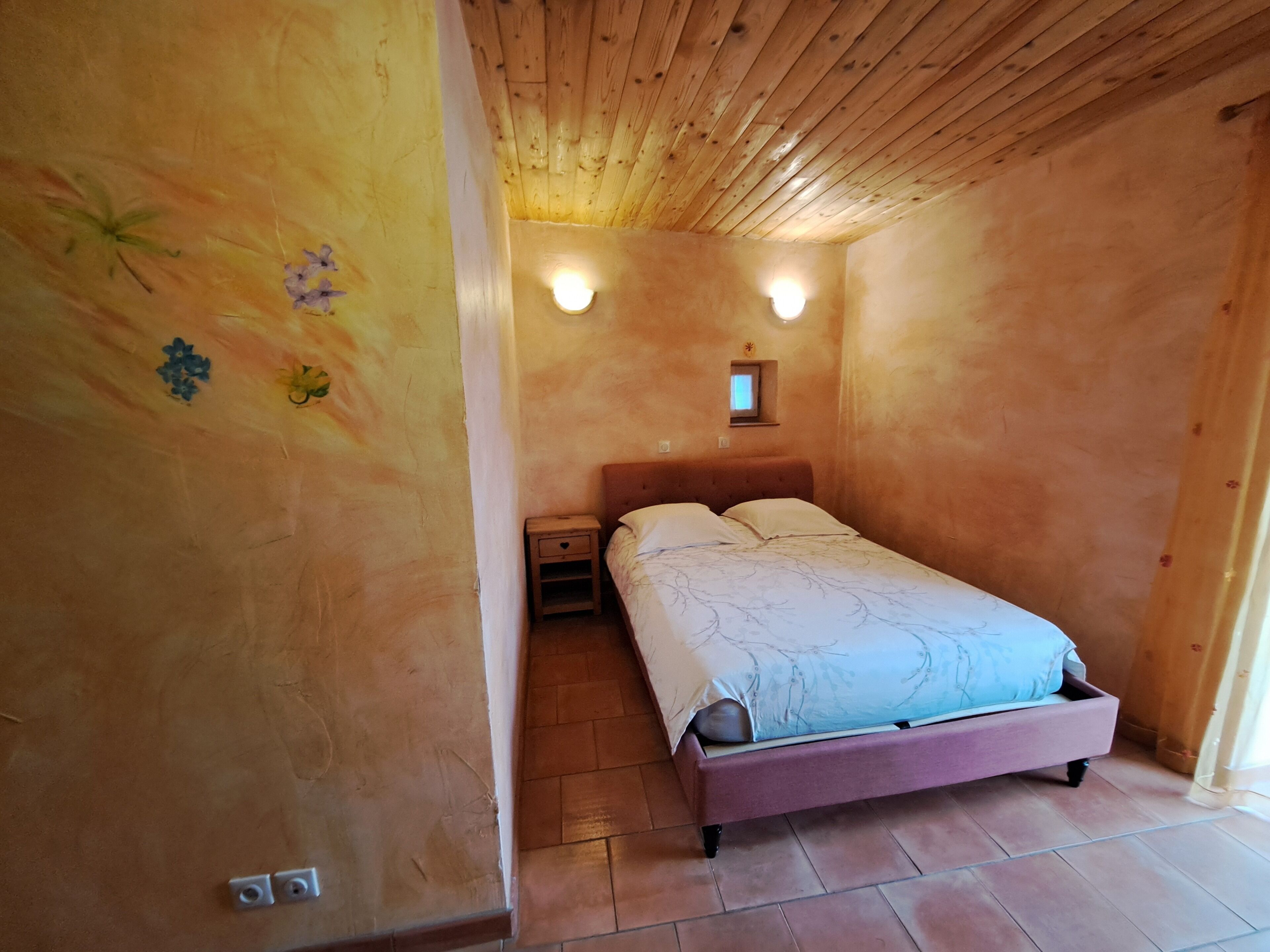 A bedroom with a wooden ceiling, a bed with a headboard, two bedside tables, and a small window with a light fixture.