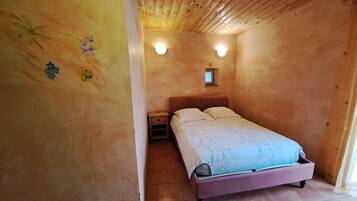 A bedroom with a wooden ceiling, a bed with a headboard, two bedside tables, and a small window with a light fixture.