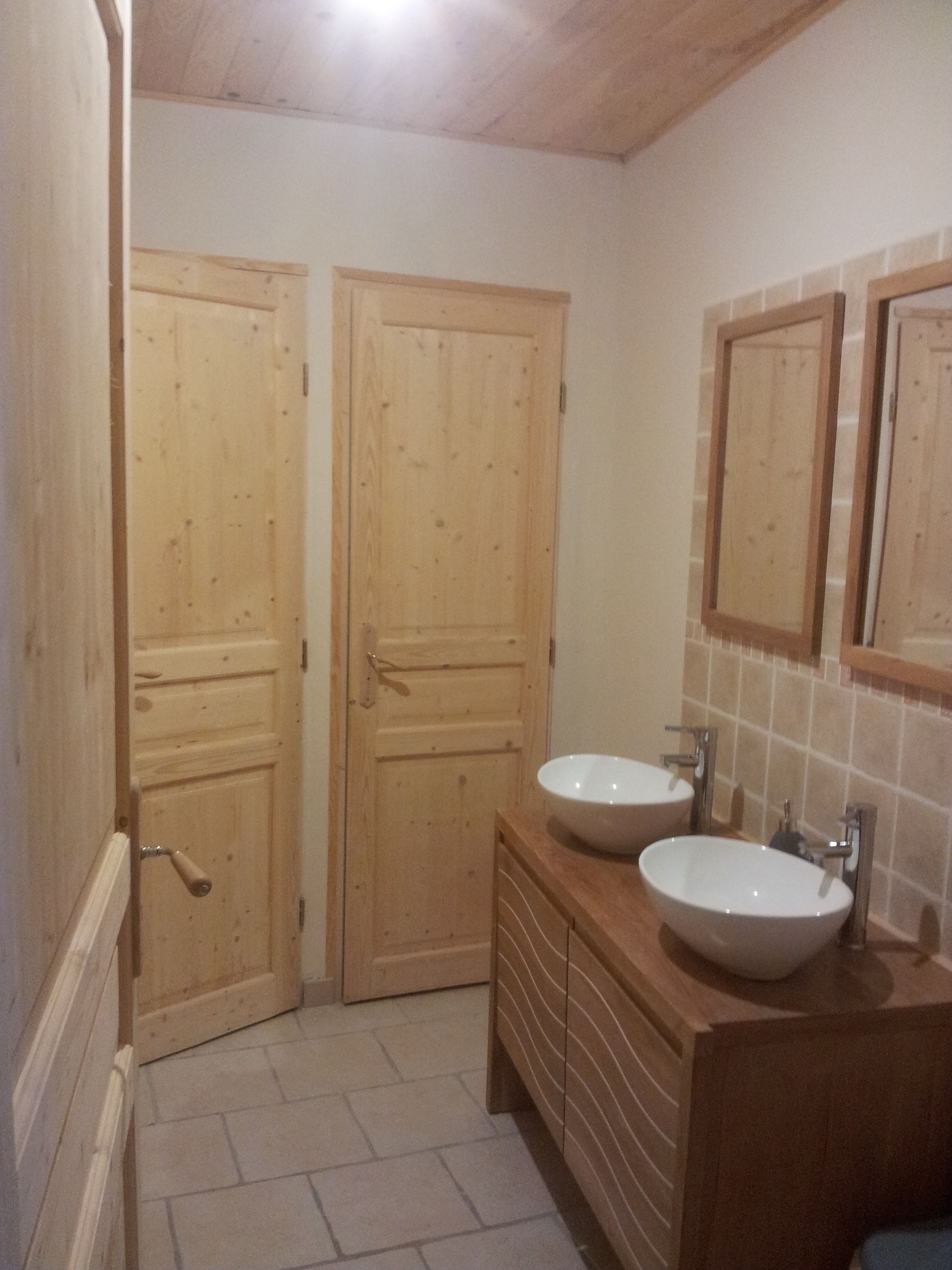 Un baño con dos puertas de madera, un tocador de madera con lavamanos y una pared con azulejos.