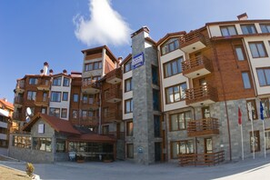 A multi-story hotel with balconies, a central stone pillar, and a small building with a red roof. - Grand Montana (Bansko)