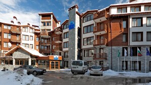 A multi-story hotel with balconies, a central pillar with signage, and a parking area with snow. - Grand Montana (Bansko)