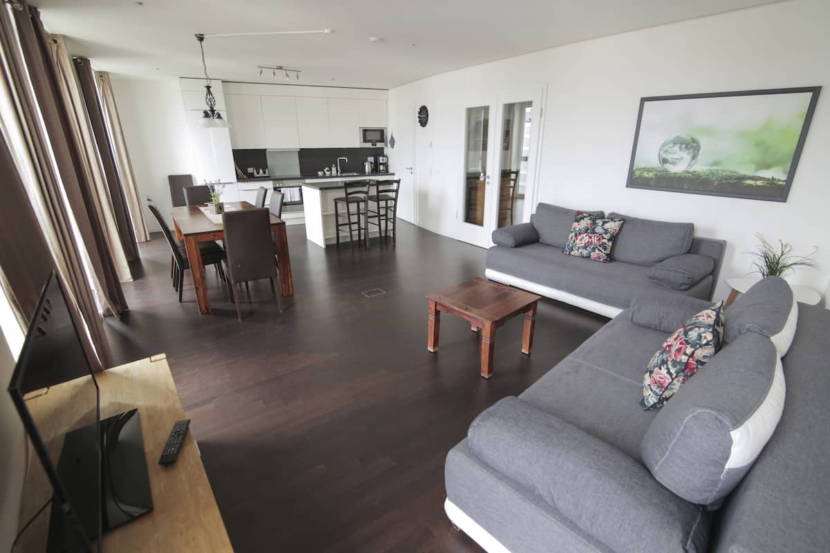Ein modernes Wohnzimmer mit grauem Sofa, einem Holztisch und einem Essbereich mit Stühlen. Im Hintergrund ist eine Küche zu sehen.