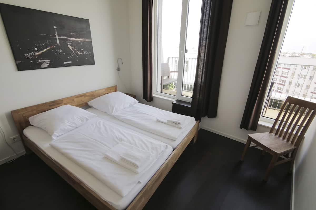 Ein Hotelzimmer mit Doppelbett, Holz-Kopfteil, einem Fenster mit Vorhängen, einem Sessel und einem Stadtbild an der Wand.