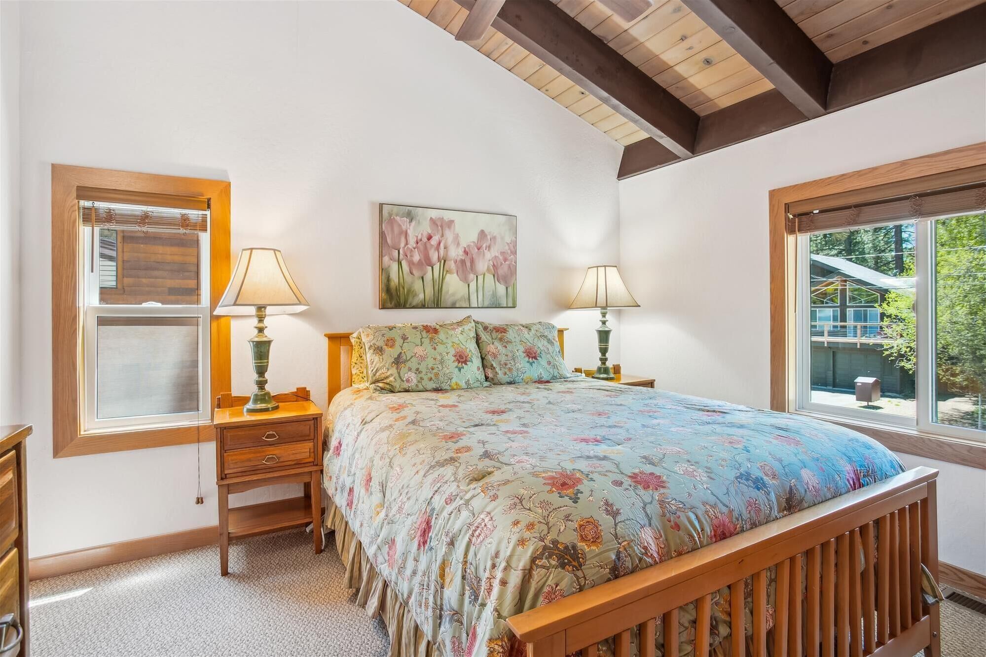 A bedroom with a wooden bed, floral bedding, two bedside lamps, a nightstand, a wooden dresser, a window with blinds, and a painting of flowers on the wall.