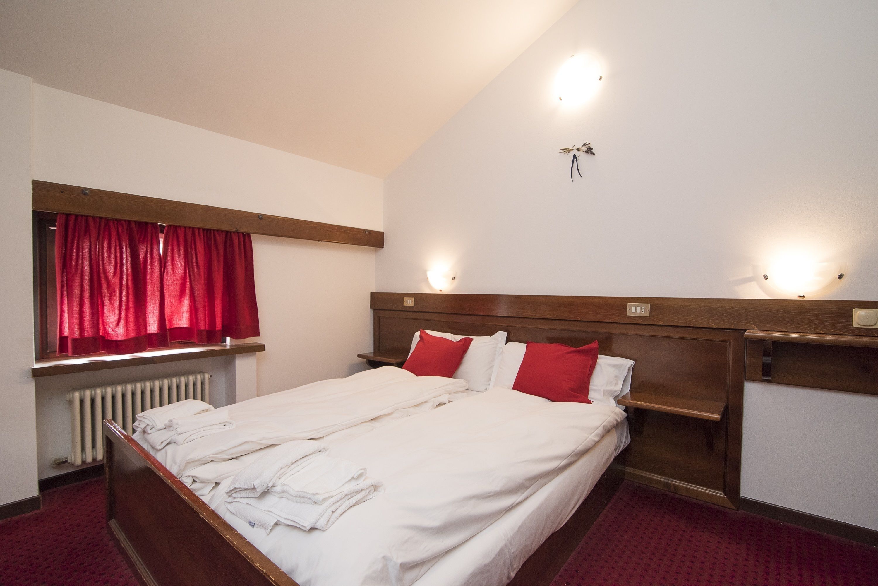 Habitación de hotel con cama, cortinas rojas, cabecera de madera, mesitas de noche e iluminación en la pared.