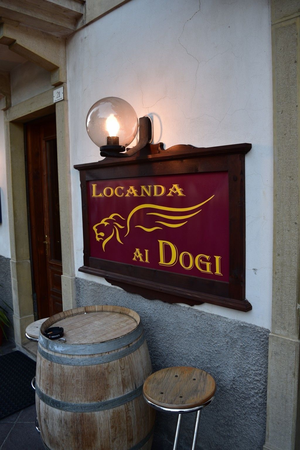 A sign for Locanda Al Dogi with a lamp above it, wooden barrels, and a stool.