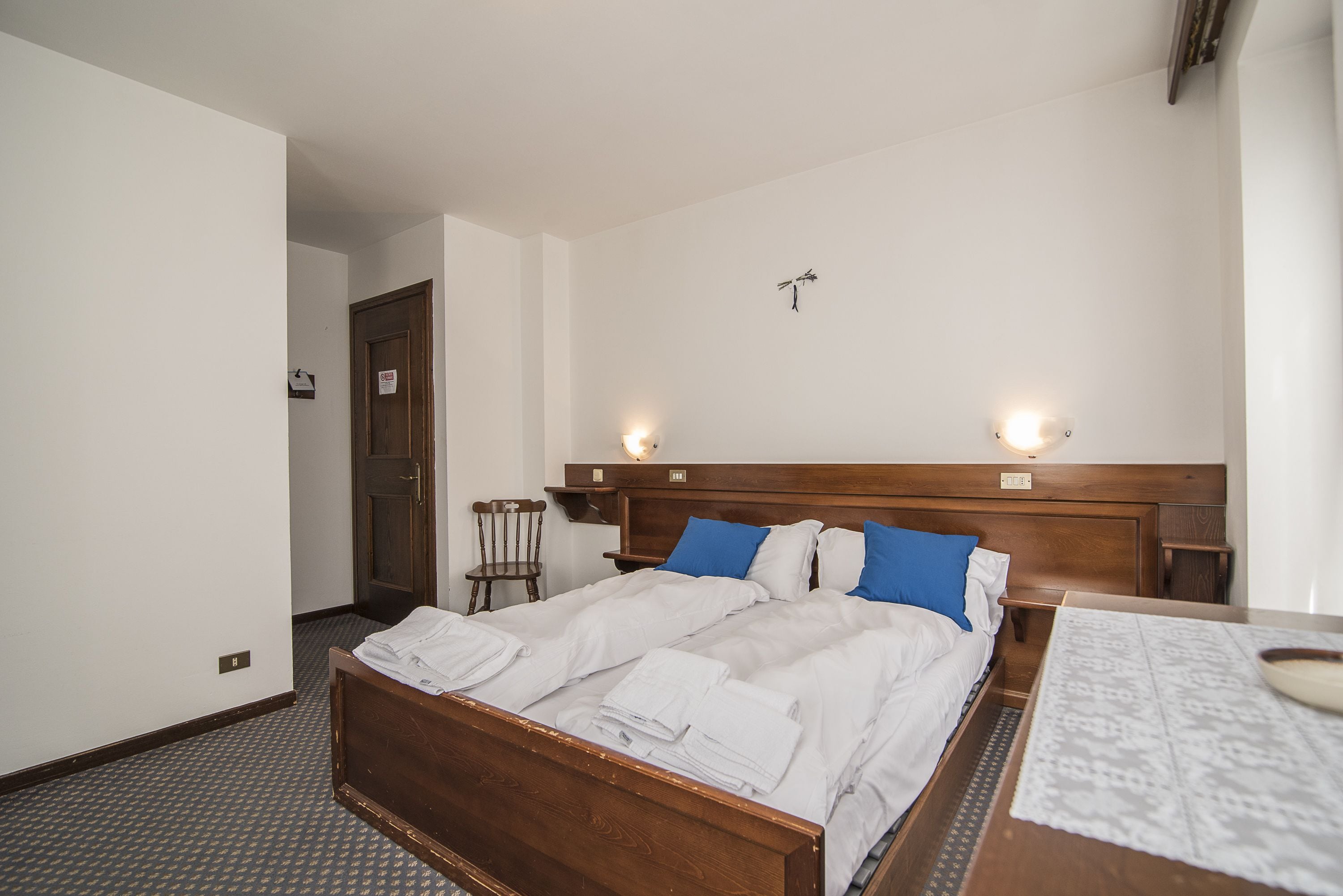 Une chambre d’hôtel avec un lit en bois, du linge de lit blanc, une table de chevet et un luminaire fixé au mur.