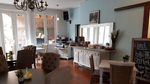 A dining area with a round table, several chairs, a kitchen area with a chalkboard menu, and a view through large windows. - Cafe pension The Chandelier (Terneuzen)
