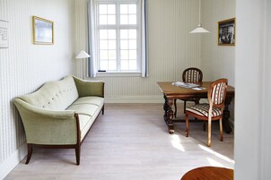 A room with a green sofa, a wooden dining table with chairs, a window with curtains, and wall-mounted lamps.
