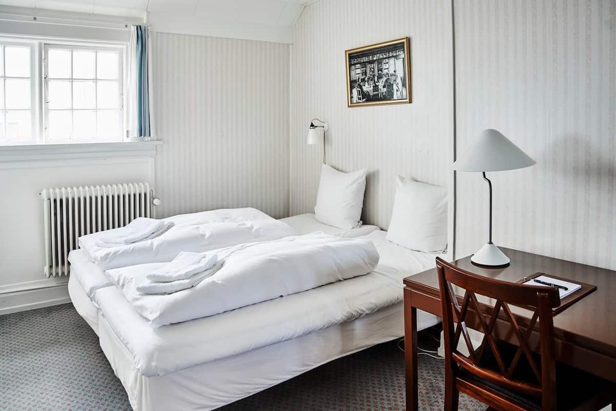 Et hotelværelse med seng, skrivebord, stol, radiator, et vindue med gardiner og et billede i ramme på væggen.