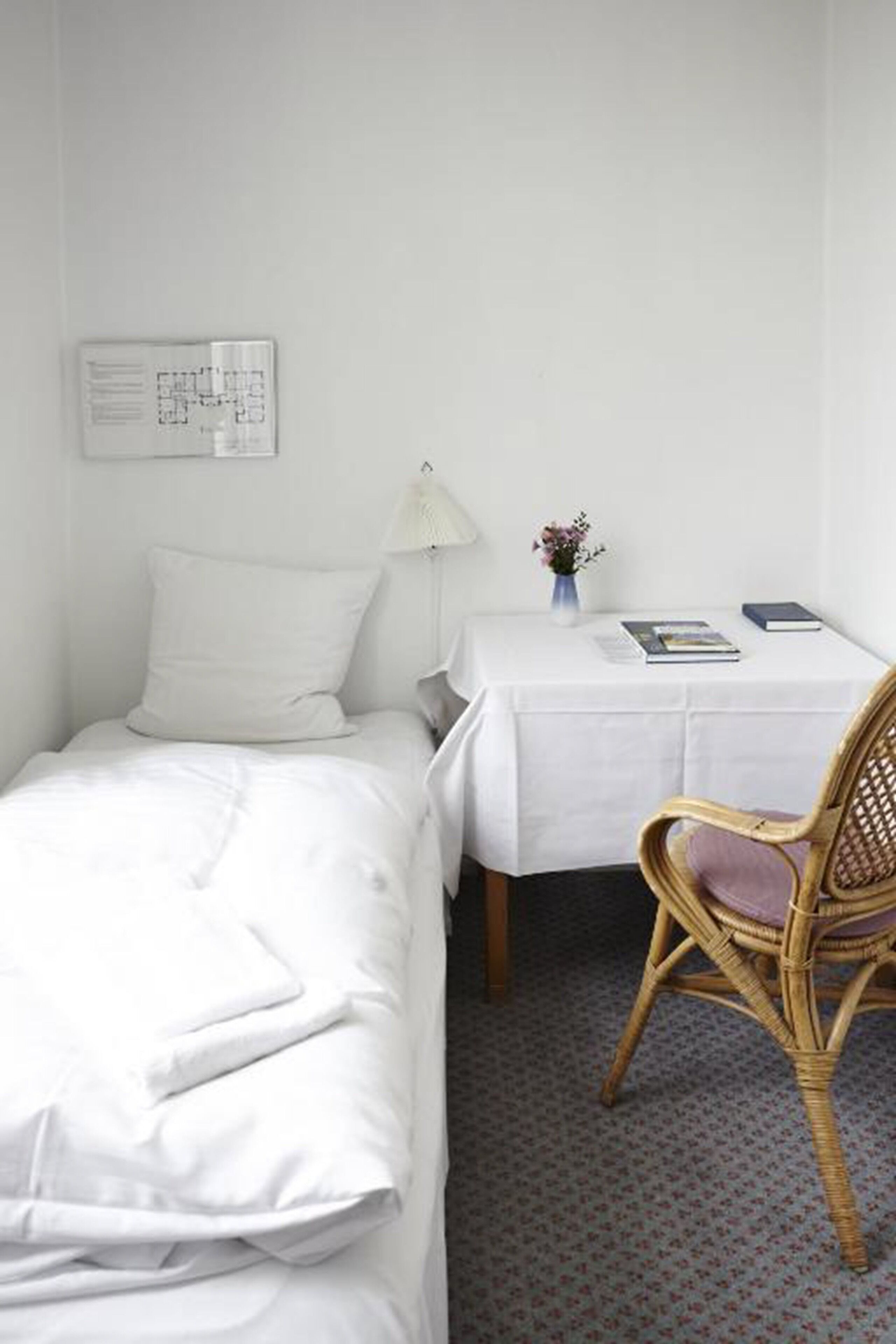 Une petite pièce, bien rangée, avec un lit simple, une petite table et une chaise, une lampe et un calendrier accroché au mur.