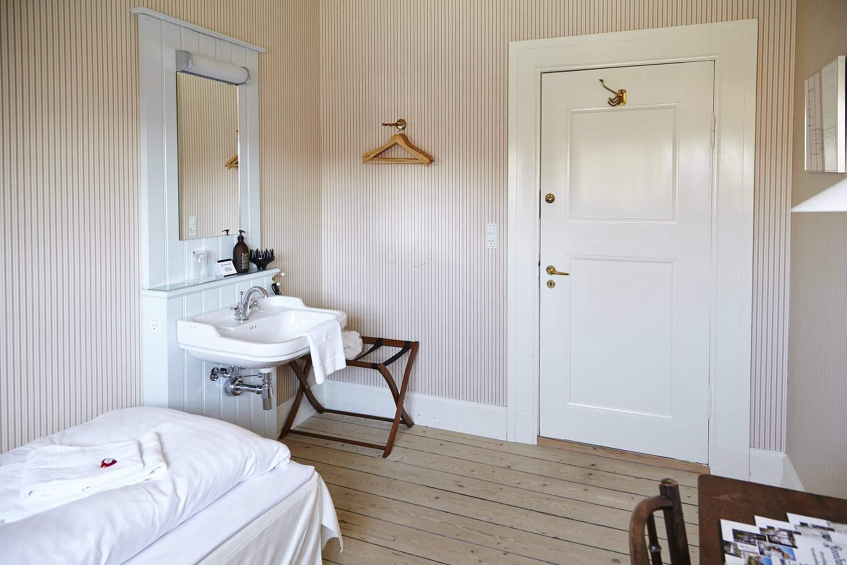 A bedroom with a bed, a bedside table, a hanging clothes rack, a white door, and a bathroom with a sink and mirror.