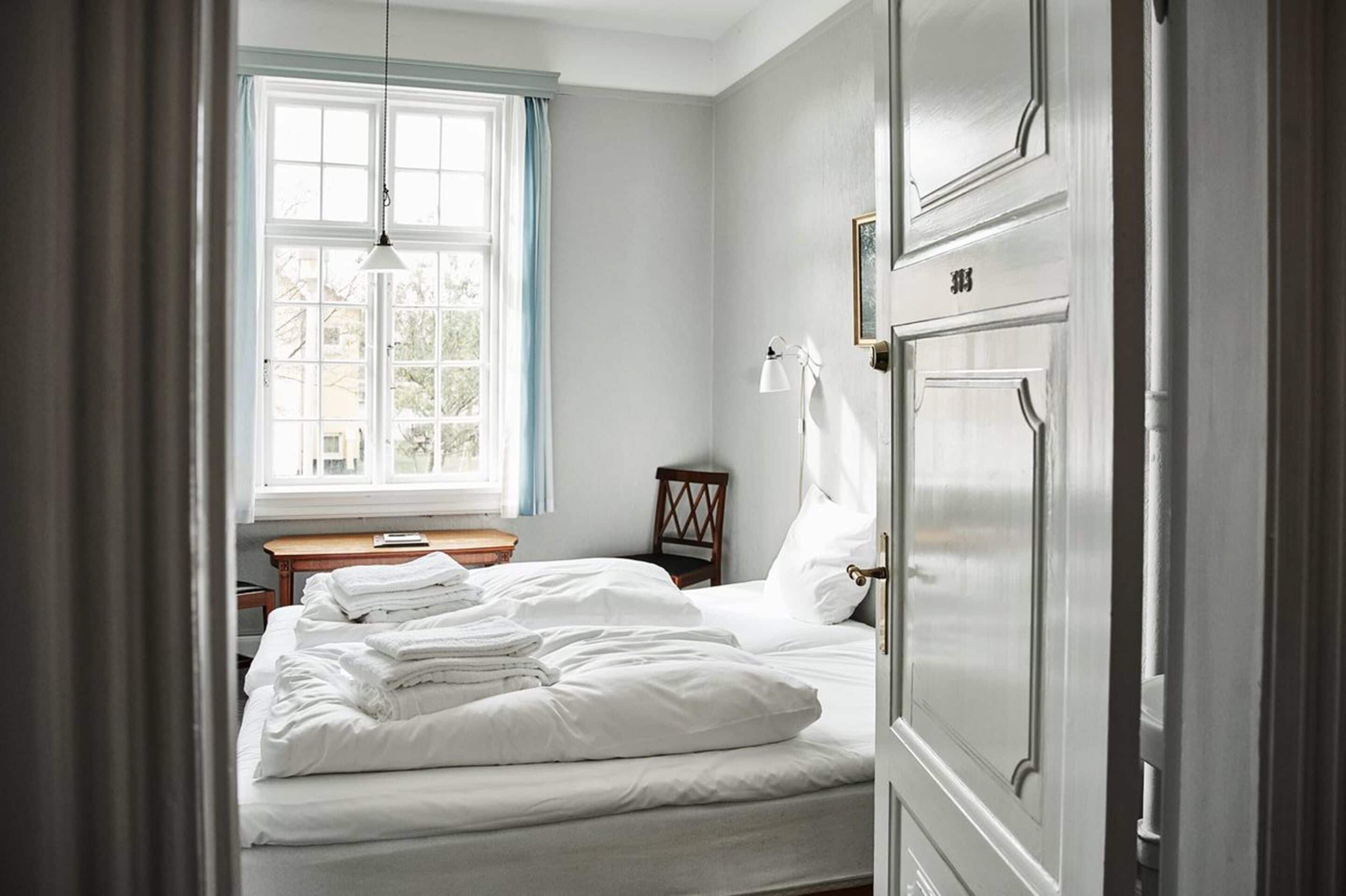 A bedroom with a bed, a nightstand, a chair, a window with curtains, and a door.