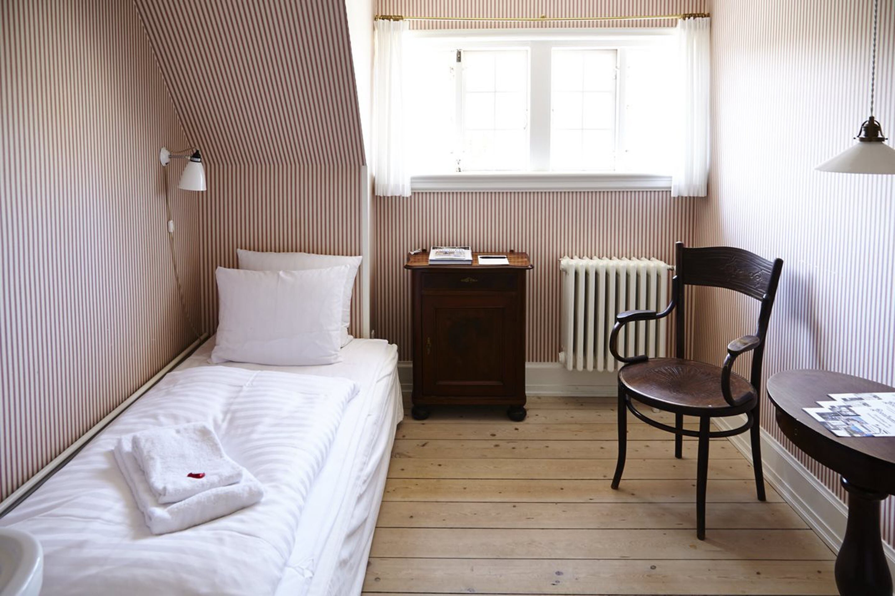 A small, neatly arranged room with a single bed, a chair, a small table, and a window with curtains.