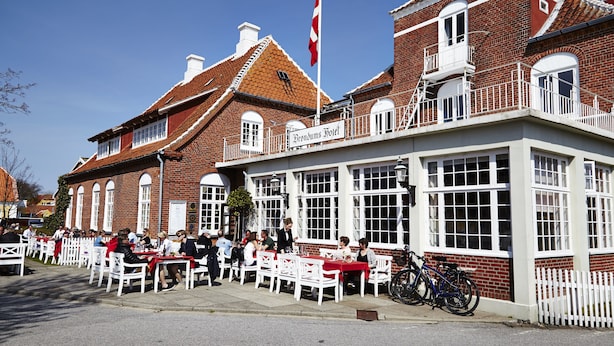 A building with a sign reading "Frondum Hotel" and a flagpole with a flag. There are outdoor tables and chairs, and bicycles parked outside. - Brøndums Hotel (Skagen)
