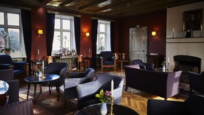 A well-lit lounge area with multiple seating options, including sofas and armchairs, arranged around a central coffee table. There are two windows with curtains, a fireplace, and a clock on the wall. - Brøndums Hotel (Skagen)