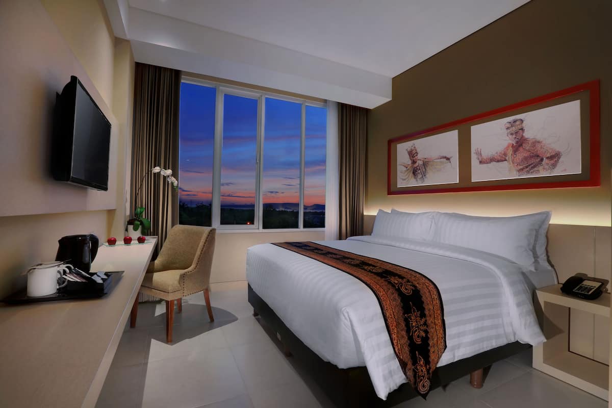A hotel room with a large bed, a desk with a coffee maker, a chair, a TV, and a window with a view of the sunset.