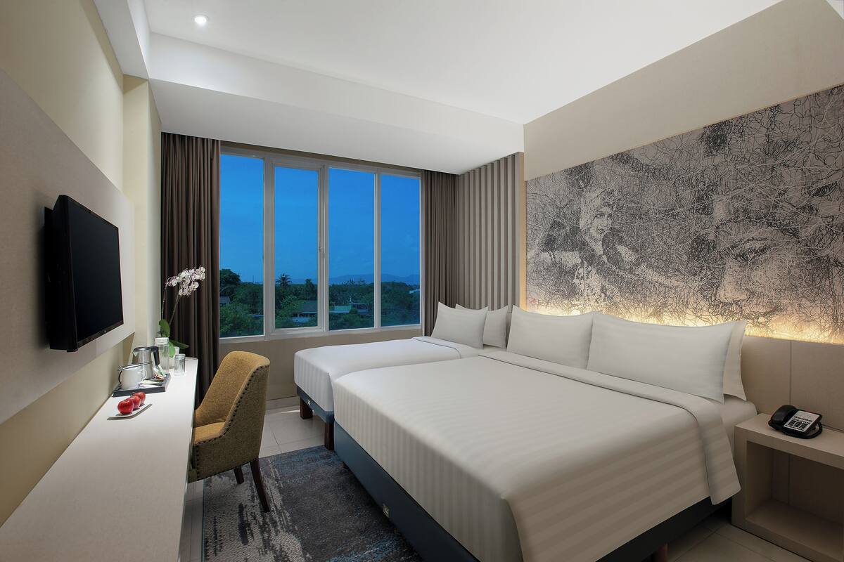 A modern hotel room with a large bed, a desk with a flat-screen TV, a chair, and a view of the city at dusk.