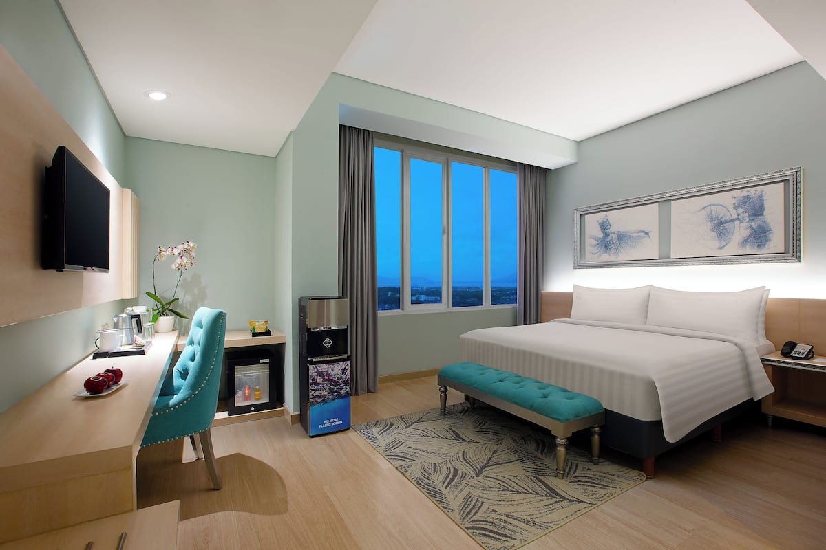 A hotel room with a bed, a desk with a chair, a TV, a window with curtains, and artwork on the wall.