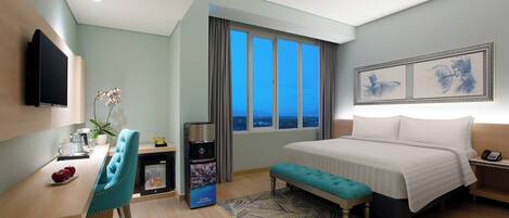 A hotel room with a bed, a desk with a chair, a TV, a window with curtains, and artwork on the wall.