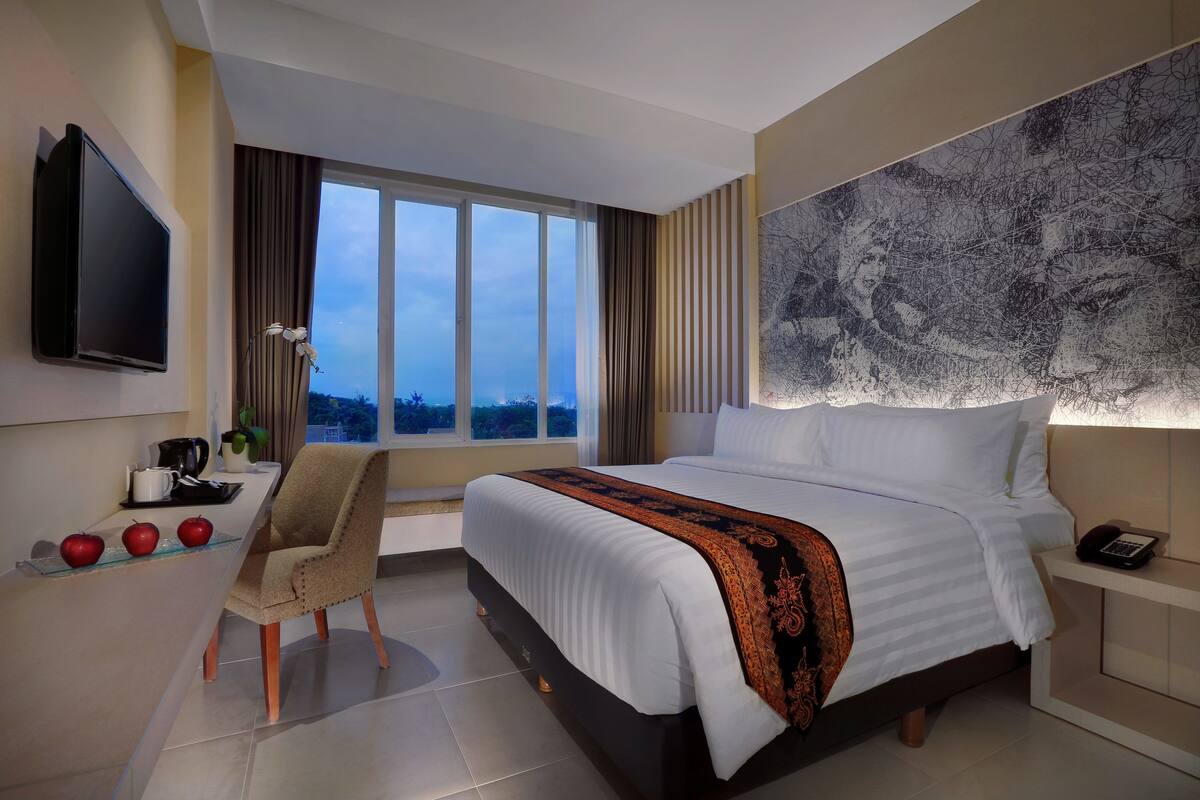 A modern hotel room with a large bed, a flat-screen TV, a desk with apples, and a view of the city.