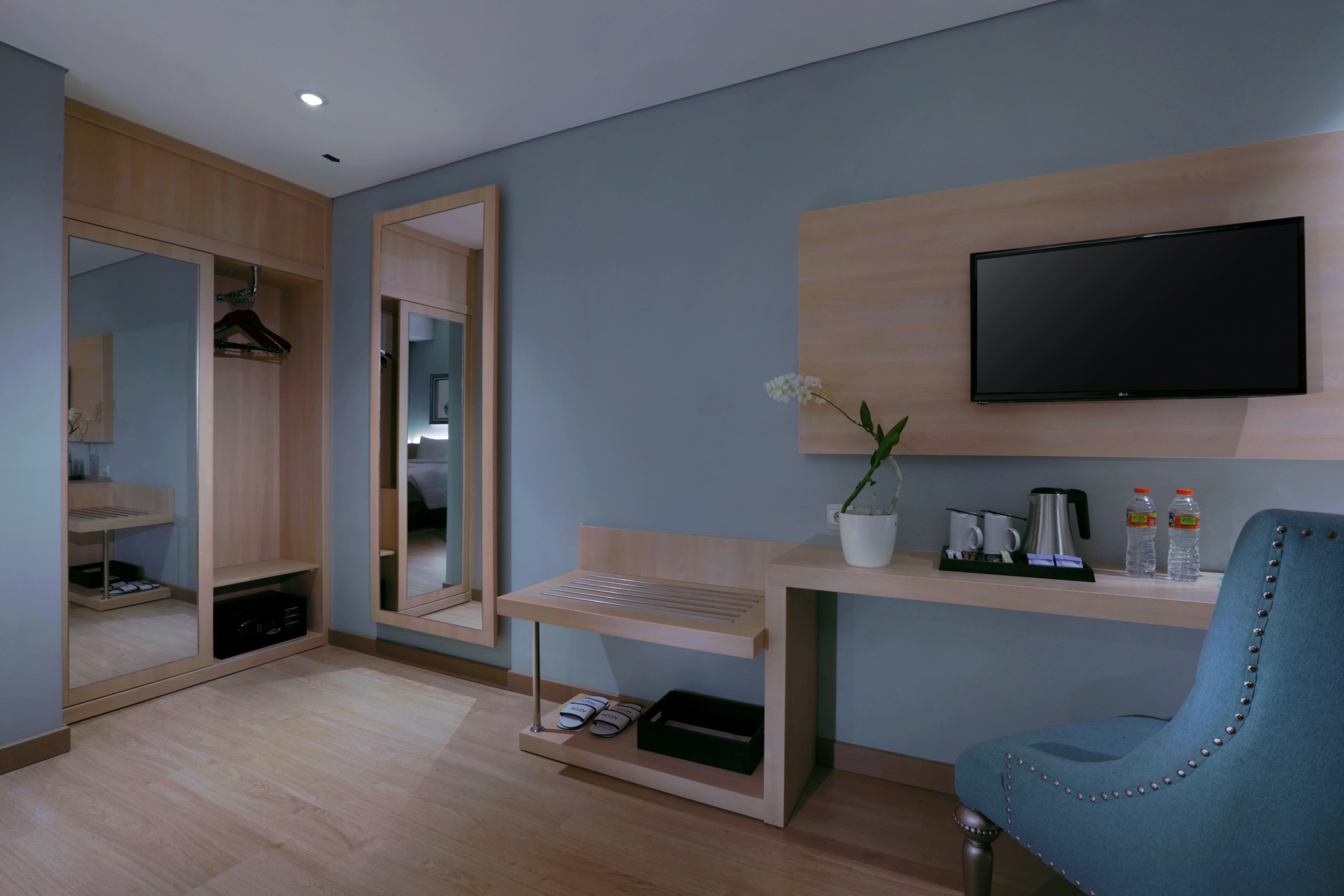 A modern hotel room with a wooden wardrobe, a mirror, a mounted TV, a desk with a chair, and a small plant.