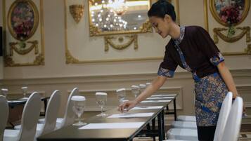 A hotel dining area with a long table set for a meal, white chairs, and a waiter serving.
