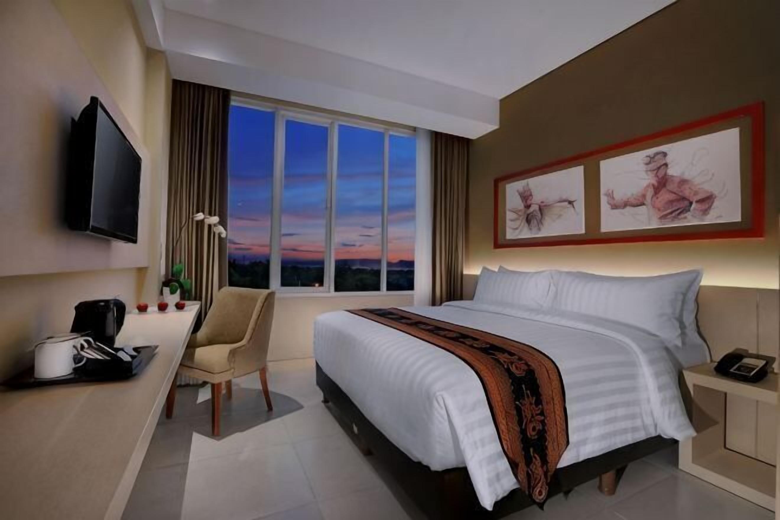 Ein Hotelzimmer mit Bett, Schreibtisch mit Stuhl, Fernseher und Kunstwerk an der Wand.