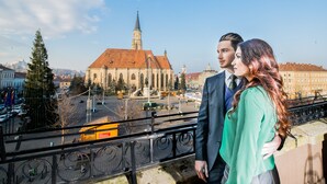 A couple standing on a balcony overlooking a cityscape with a prominent church and a tram in the background. - Red Hotel Downtown Unirii (Cluj-Napoca)