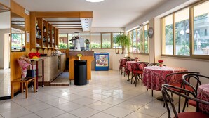 A restaurant with a counter, shelves with bottles, a trash bin, and tables with red chairs. - Hotel Levante (Cervia)