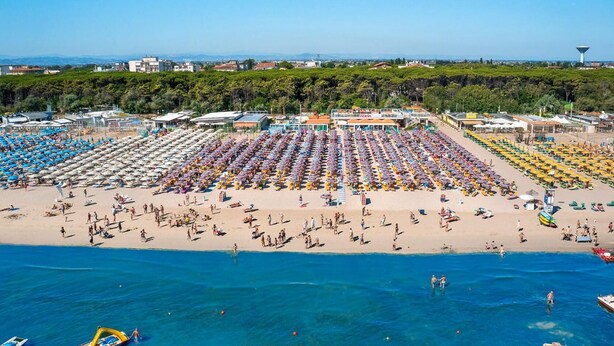 A beach with rows of umbrellas and sun loungers, a clear blue sea, and buildings in the background. - Hotel Levante (Cervia)