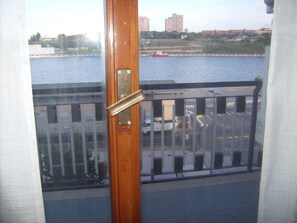 Uitzicht vanuit een raam met een houten deur, een metalen hendel en een glazen balkonhek op een waterpartij en gebouwen.