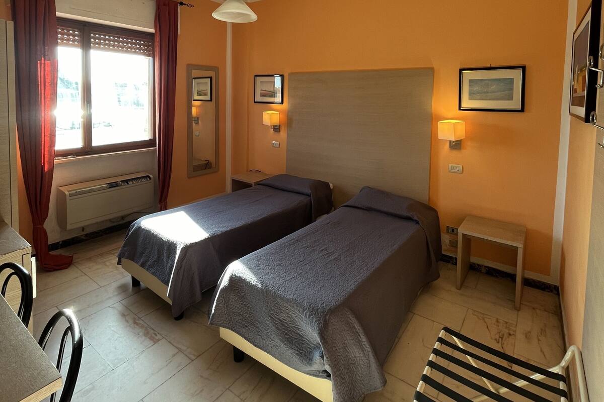 A hotel room with two single beds, a desk, a chair, a small table, and a window with curtains.