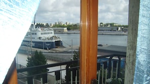 Uitzicht vanaf een balkon over een haven met een groot schip in de haven, een stadsgezicht op de achtergrond en een bloempot op het balkon.