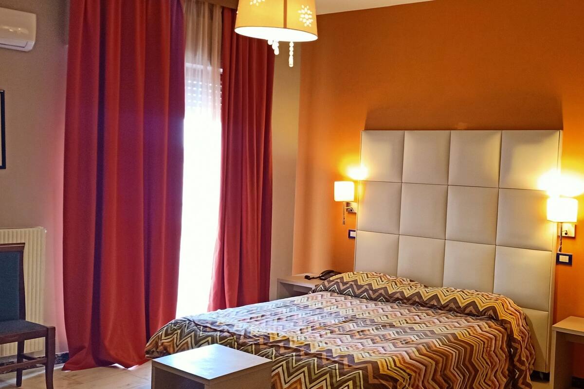 A hotel room with a large bed, two bedside lamps, a nightstand, a chair, and a window with red curtains.