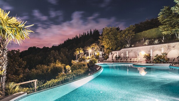 A swimming pool with a curved edge, surrounded by lush greenery and a stone wall. - Residence Madrigale (Costermano sul Garda)