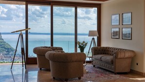 A spacious room with large windows offering a view of the sea and a distant city. There are two sofas and a tripod stand. - Residence Madrigale (Costermano sul Garda)