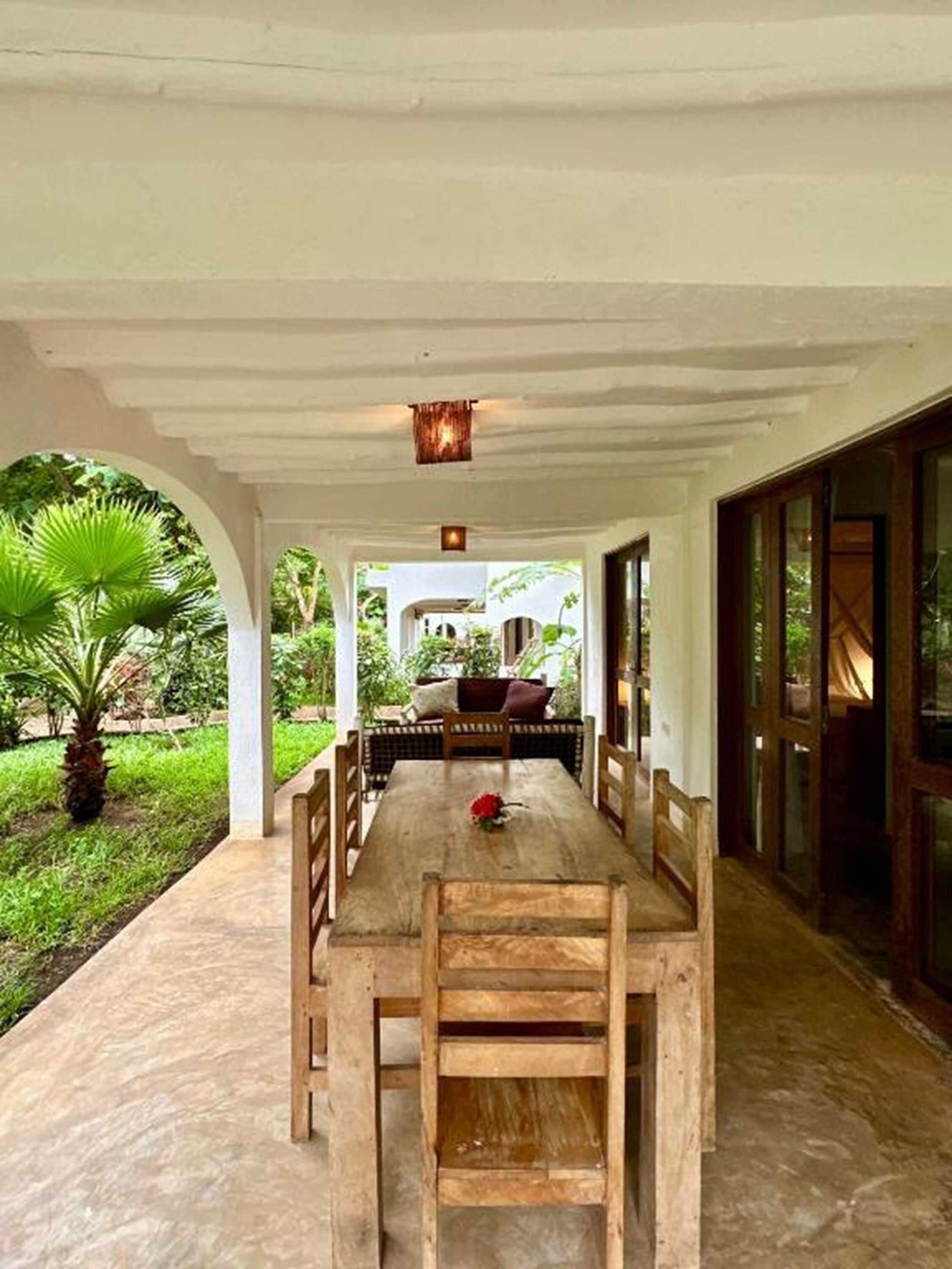 A covered outdoor dining area with a wooden table and benches, surrounded by greenery and a view of a garden.