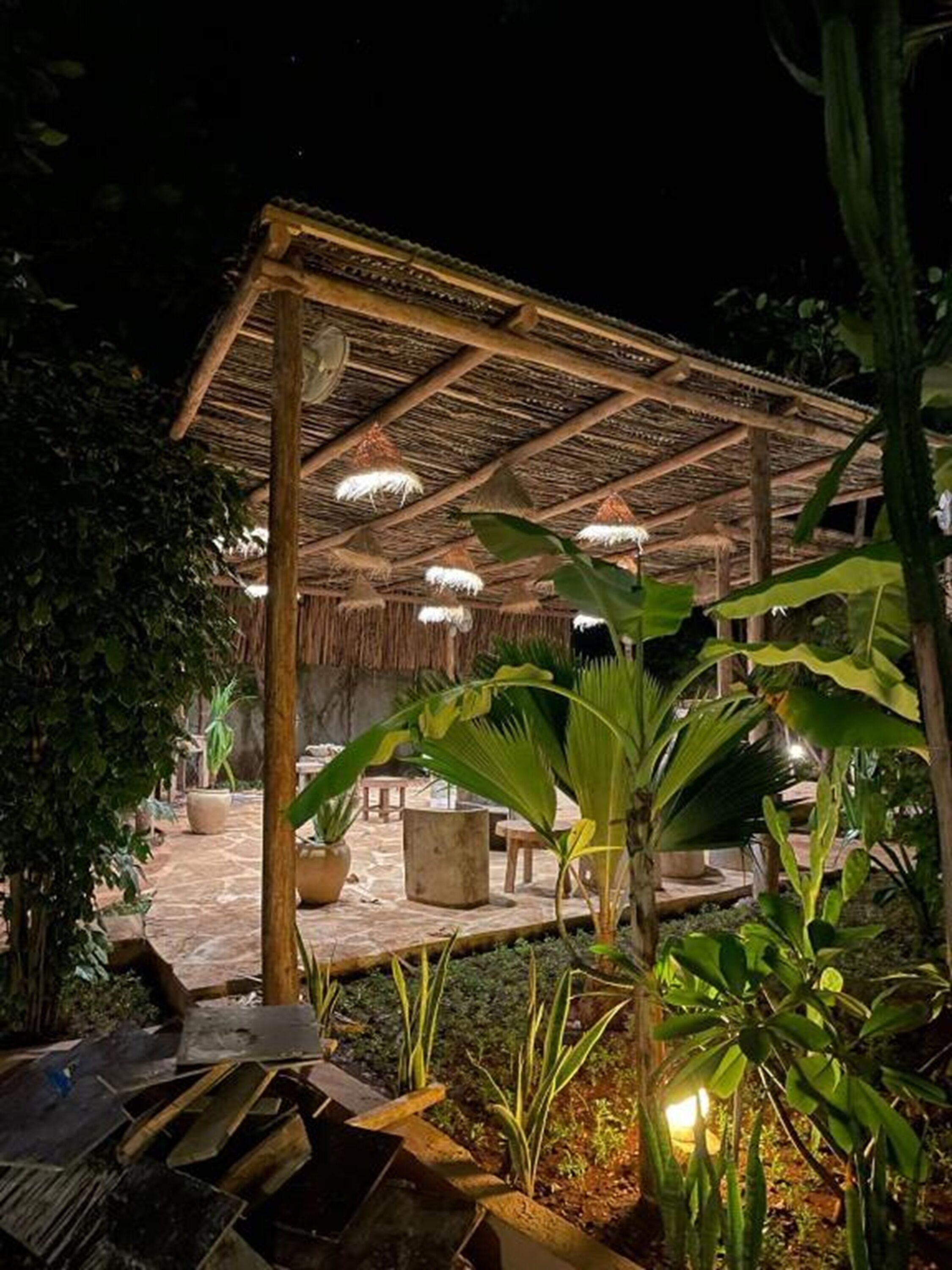 Área externa com telhado de palha, móveis de madeira e plantas em vasos.