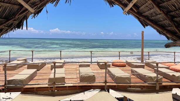 Een strandlounge met houten terras en rieten dak, met uitzicht op helder turquoise water en een zandstrand.