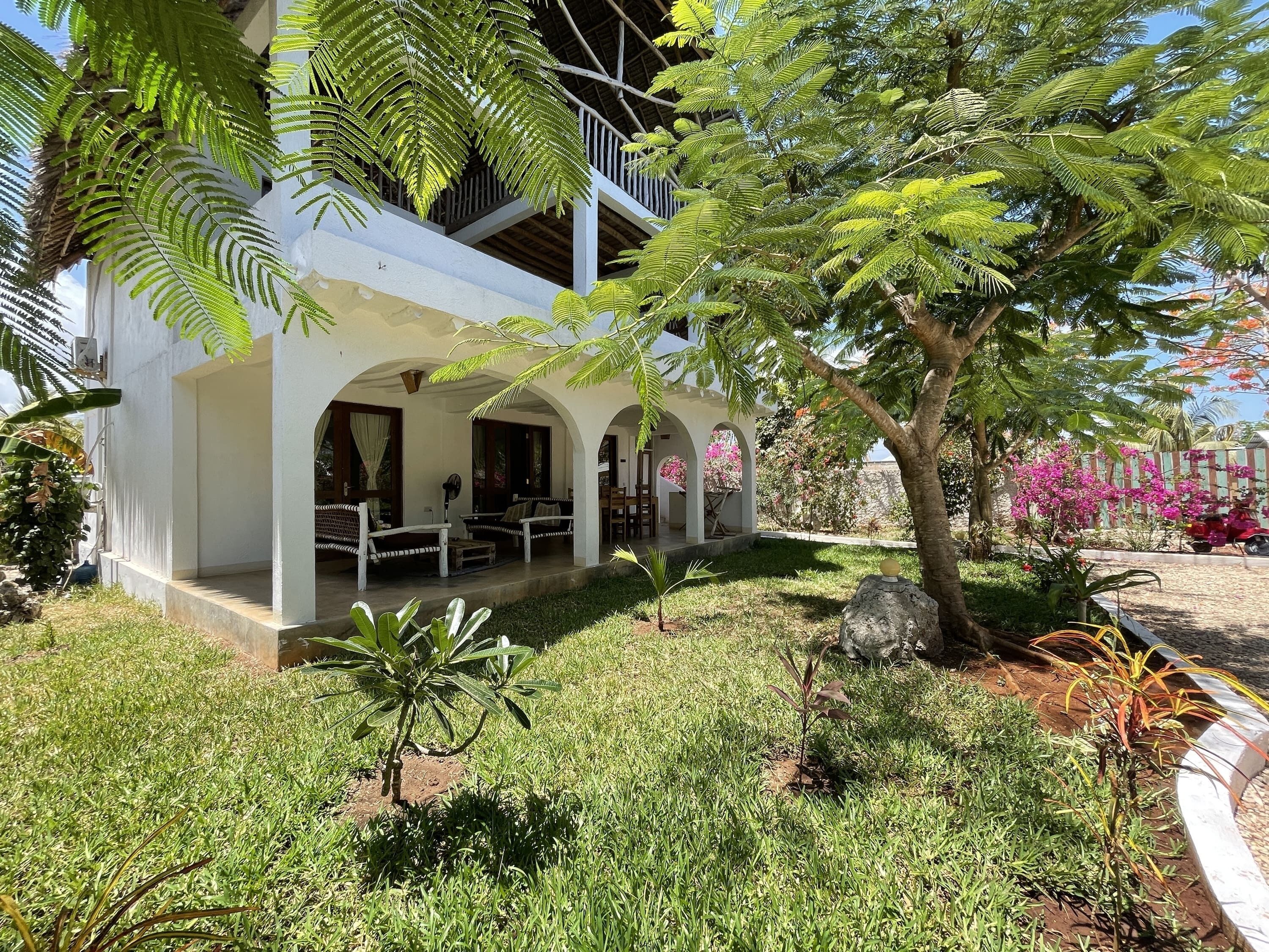 A white building with a covered porch, outdoor seating, and a well-maintained garden with various plants and a tree.
