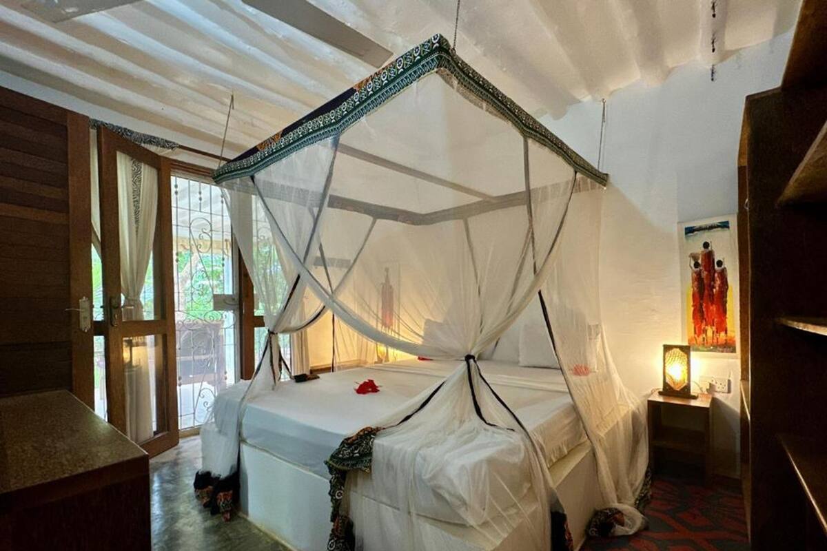 Une chambre avec un lit sous un dais, un plancher en bois, une fenêtre avec des rideaux et une lampe fixée au mur.