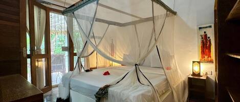 A room with a bed under a canopy, a wooden floor, a window with curtains, and a wall-mounted lamp.