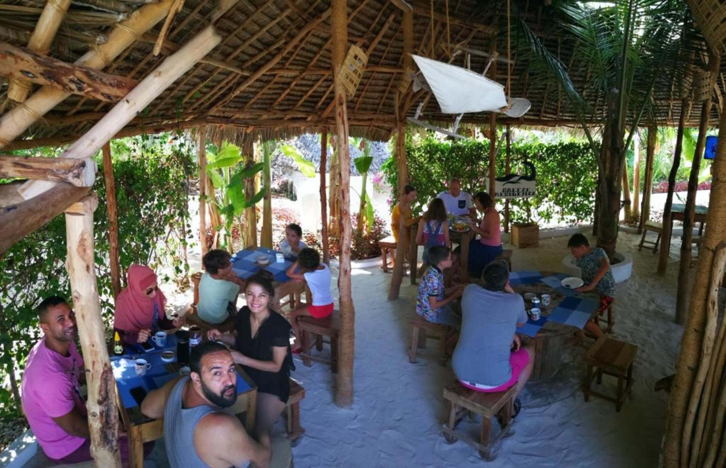 An outdoor dining area with tables and chairs under a thatched roof, surrounded by greenery.