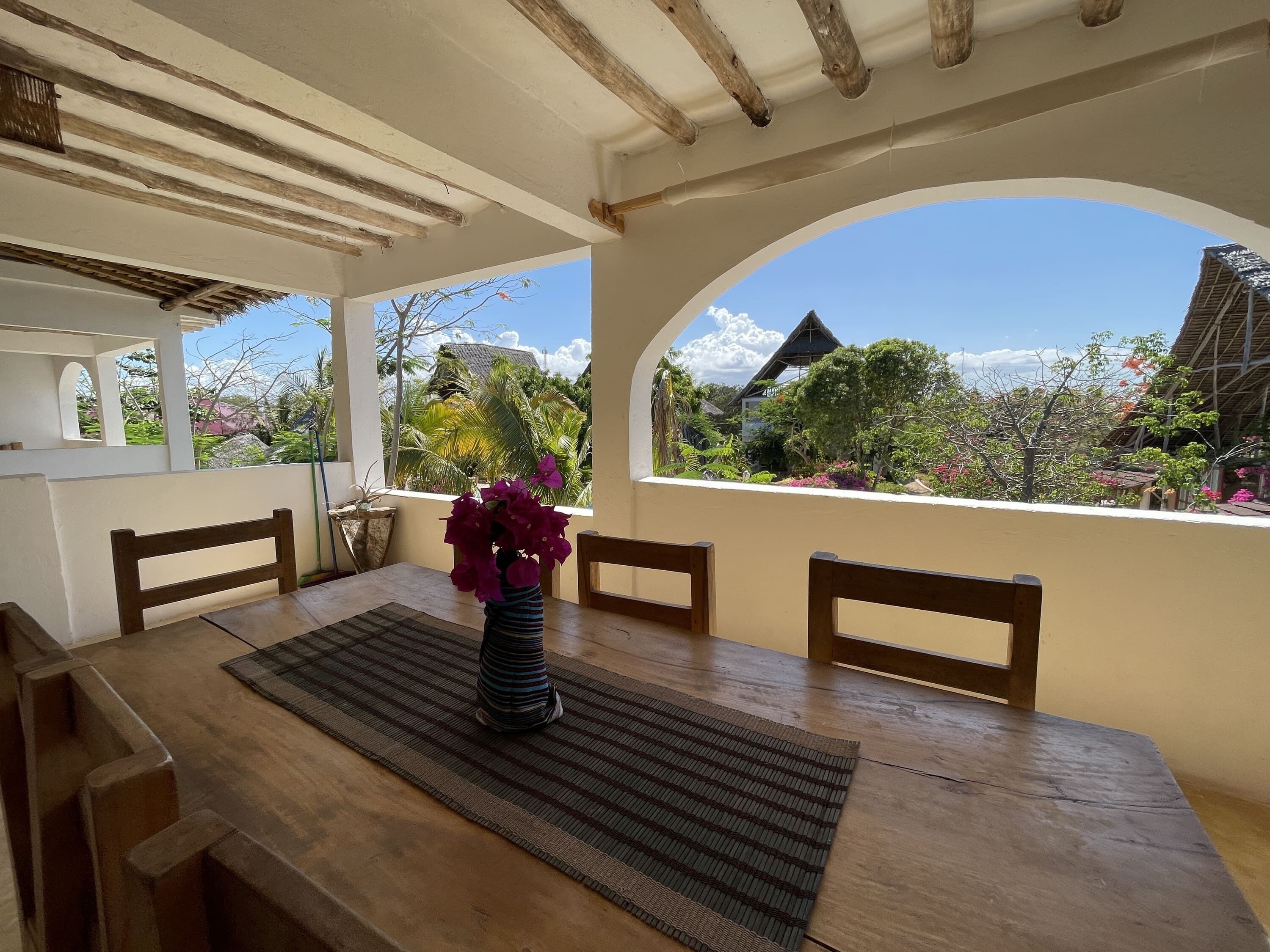 A wooden dining table with chairs, a vase with purple flowers, and a view of a tropical garden through an arched window.