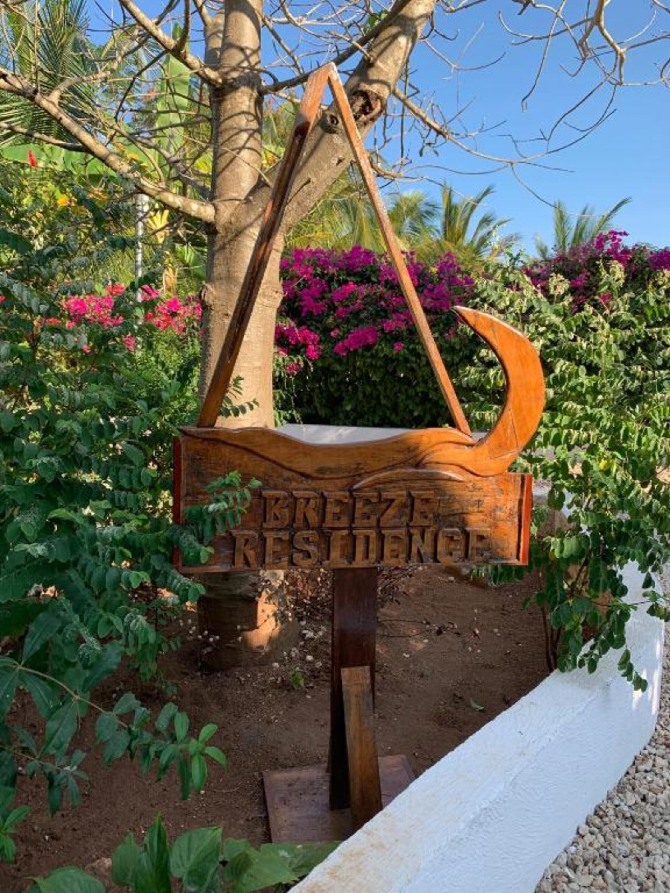 Une pancarte en bois portant l’inscription « BREEZE RESIDENCE » est suspendue à un arbre, entourée d’une végétation luxuriante et de plantes en fleurs.