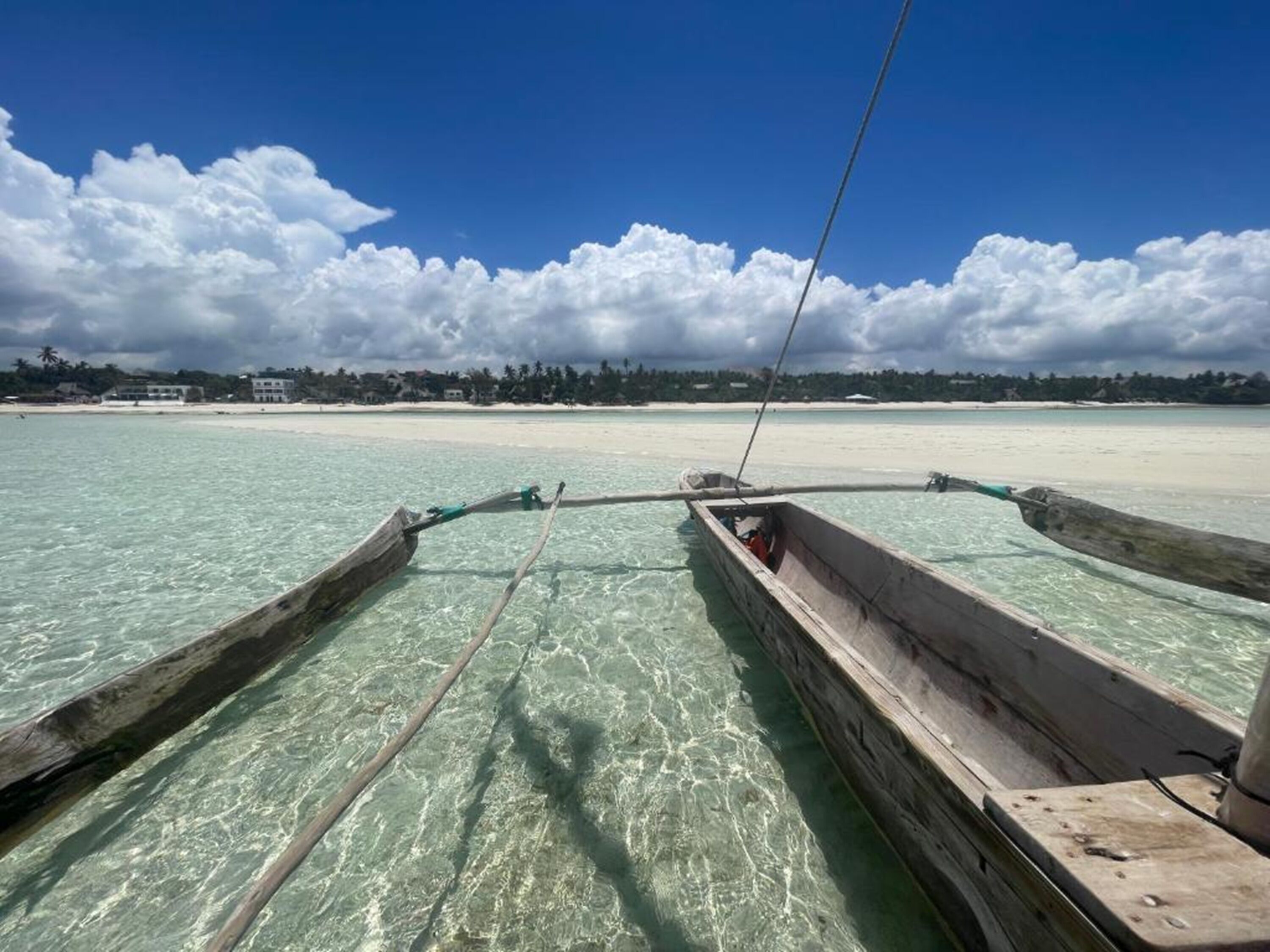 On aperçoit clairement un bateau en bois sur une eau turquoise peu profonde, avec une plage de sable et un ciel nuageux en arrière-plan.
