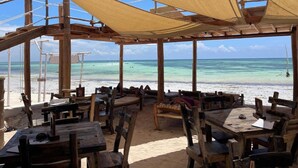 Een openluchtrestaurant aan het strand, met houten tafels en stoelen, een beschaduwd terras en uitzicht op de oceaan.